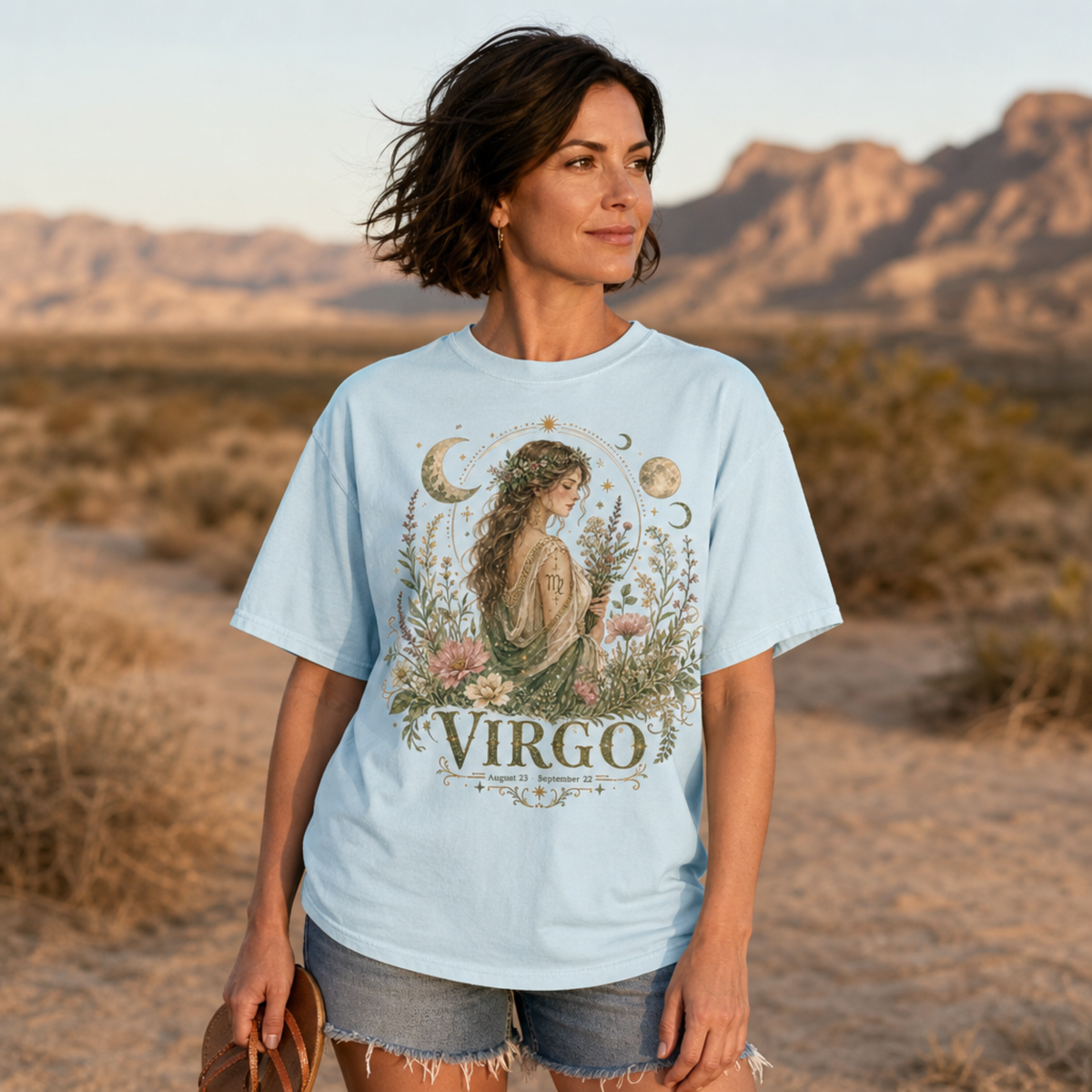 Woman wearing chambray Comfort Colors t-shirt with a mystical boho Virgo zodiac design, featuring a watercolor illustration of an ethereal woman adorned with a floral crown and surrounded by wildflowers and celestial motifs, standing in a desert landscape; perfect for astrology enthusiasts, nature and fantasy art lovers, by Crossroads Dandelion