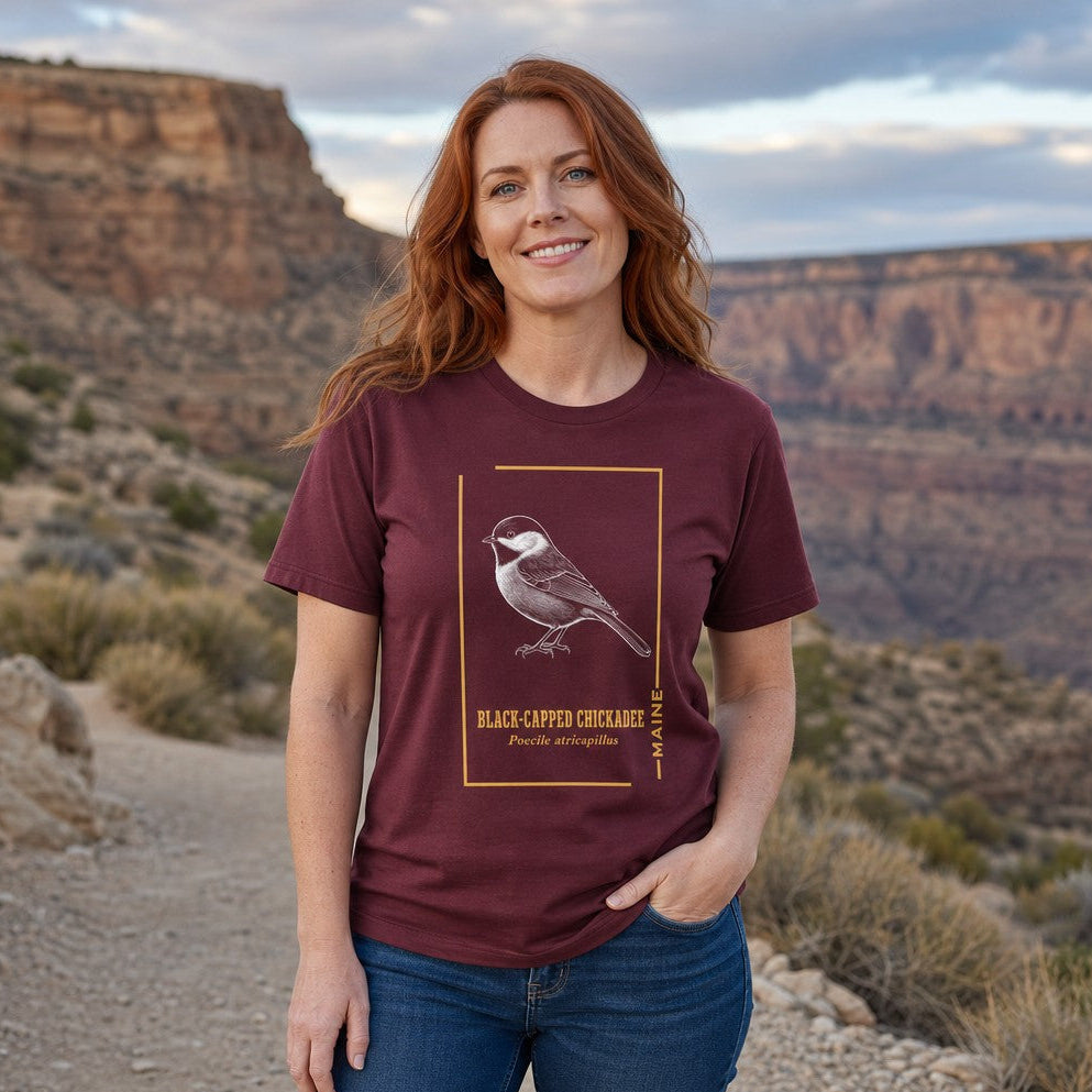 Woman wearing a maroon t-shirt featuring a detailed line art illustration of a black-capped chickadee with scientific name, vintage nature design, perfect for bird lovers and nature enthusiasts, standing on a scenic desert trail, by Crossroads Dandelion