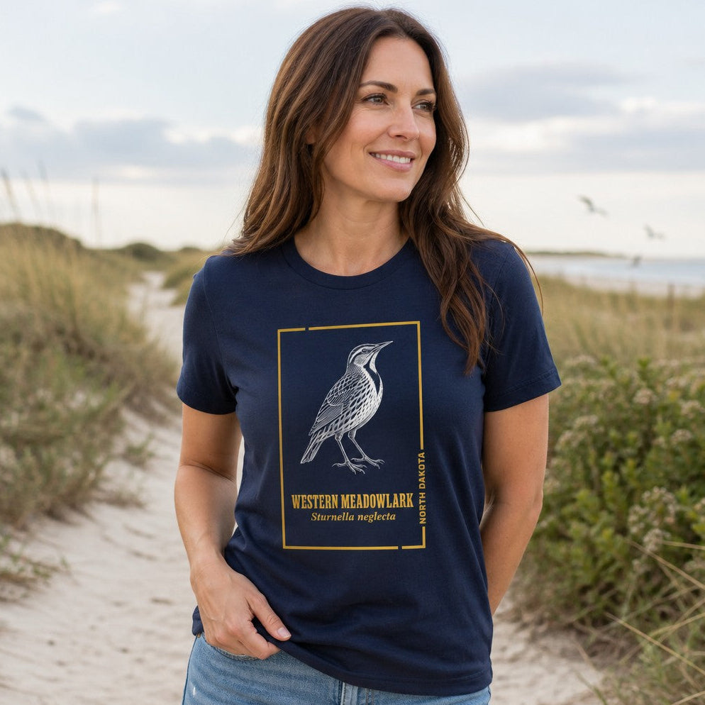 Woman wearing navy Bella Canvas 3001 t-shirt featuring intricate line art of a Western Meadowlark with vintage scientific labeling, framed in gold, standing on a sandy coastal path, heritage-inspired design for birdwatchers and nature lovers, by Crossroads Dandelion