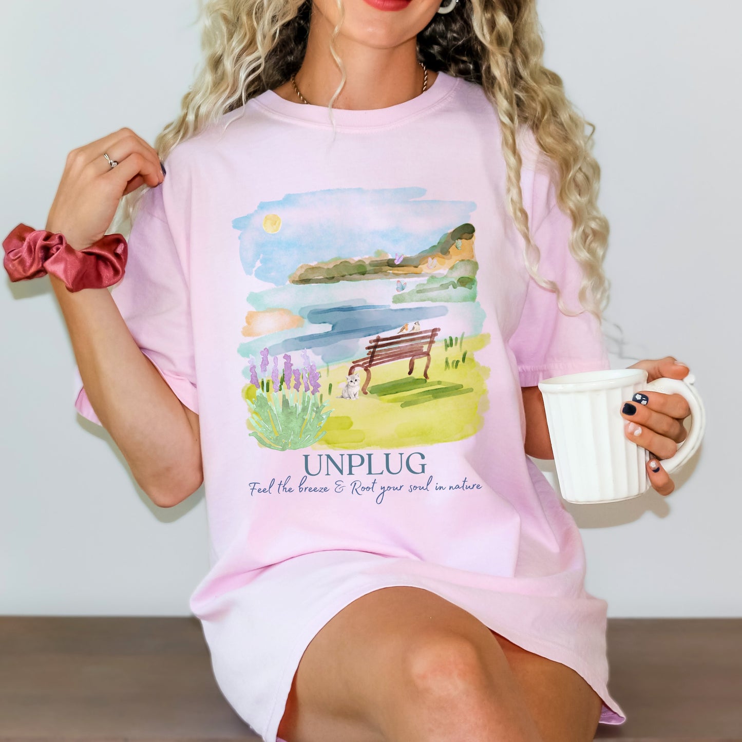 Woman wearing a Comfort Colors blossom pink t-shirt with a watercolor nature and lake graphic and 'UNPLUG' text, holding a mug by Crossroads Dandelion