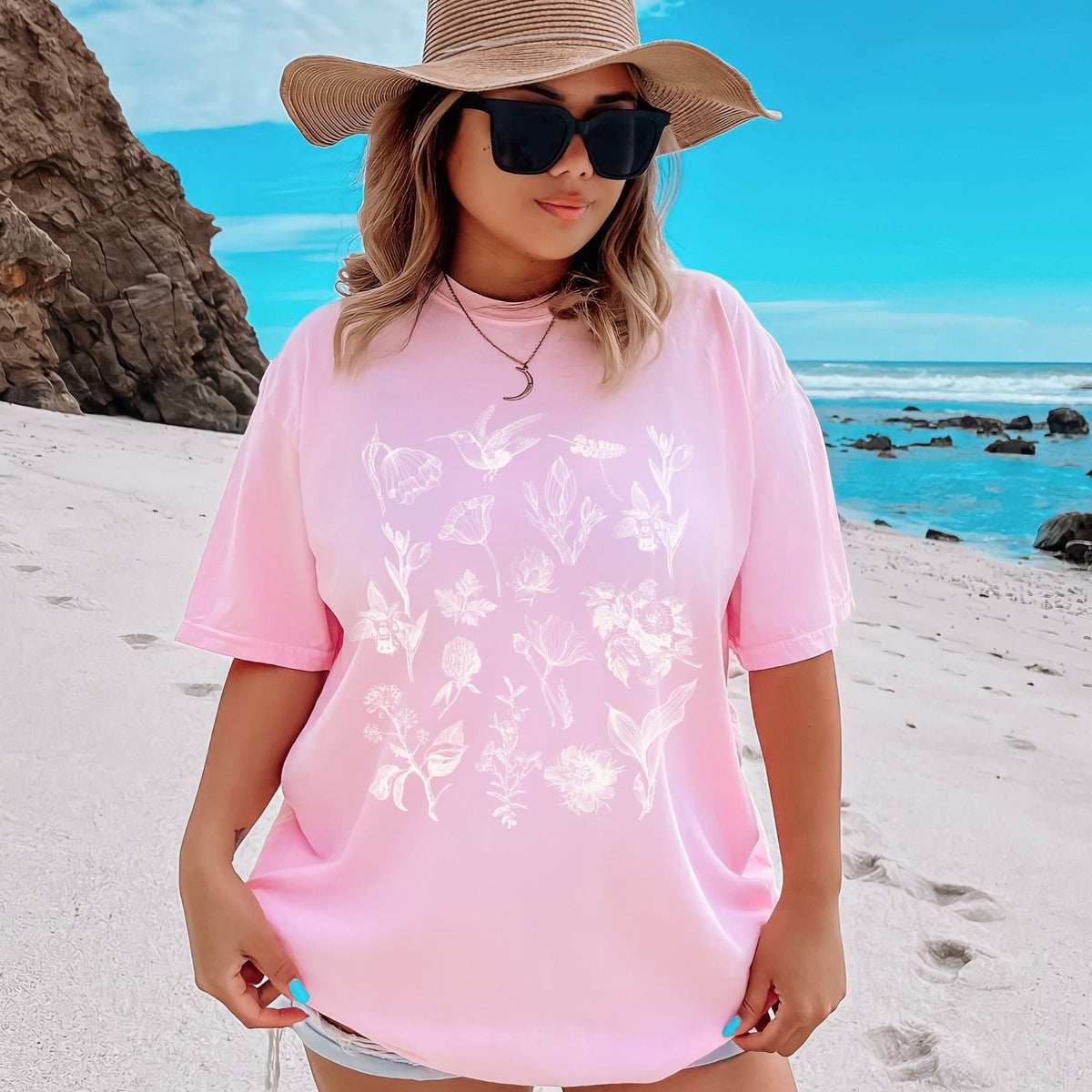 Woman in a blossom Comfort Colors floral shirt with hummingbirds and sun hat on a beach