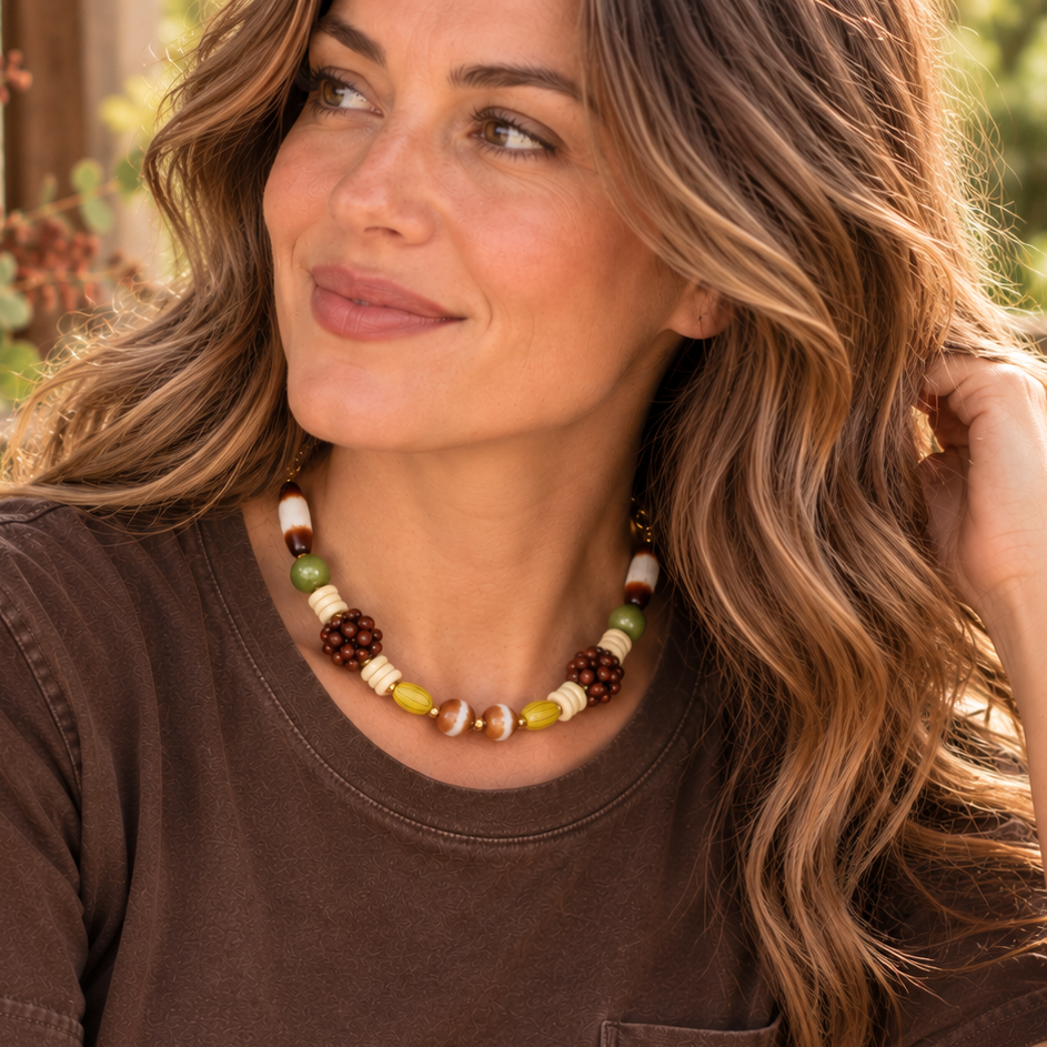 A woman with long wavy brown hair wearing a brown t-shirt and a beaded necklace with brown, green, and cream beads, sitting outdoors.