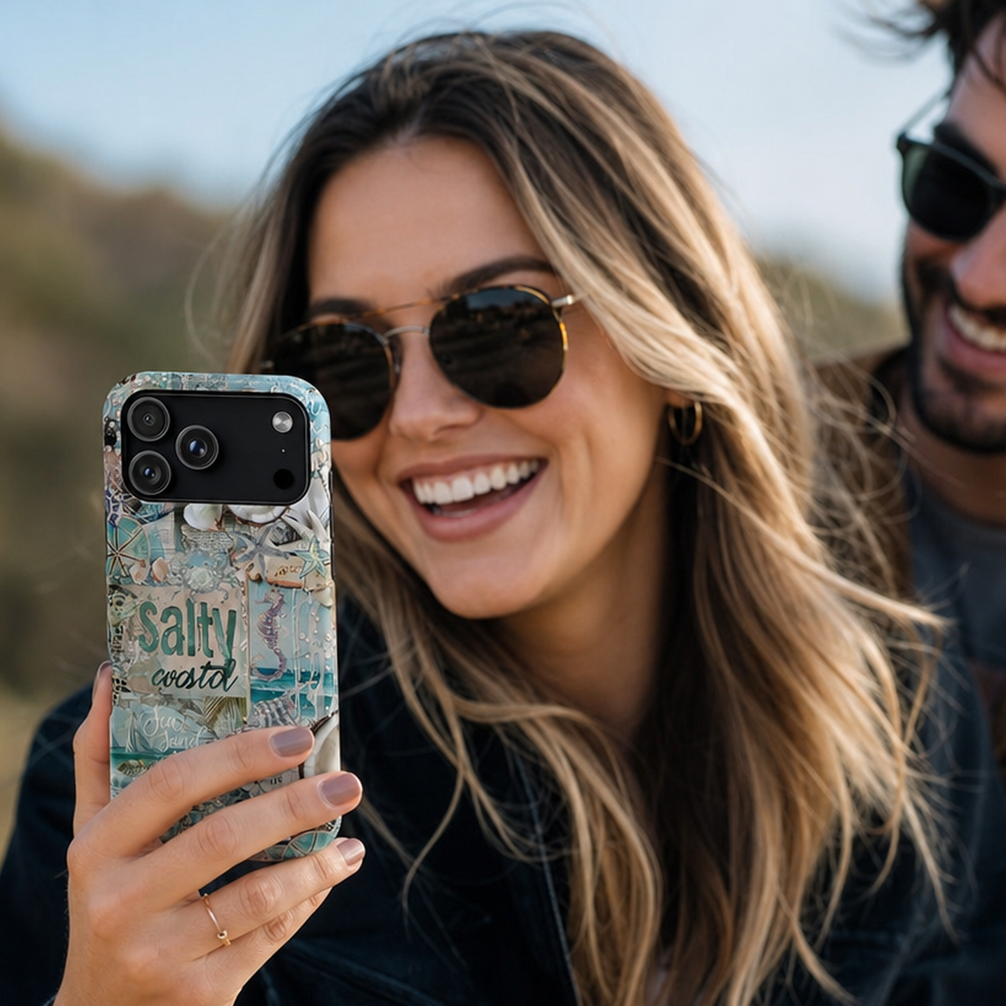 Smiling American woman in her 30s with long blonde hair wearing sunglasses using a coastal-themed iPhone 17 Pro phone case outdoors at sunset, by Crossroads Dandelion