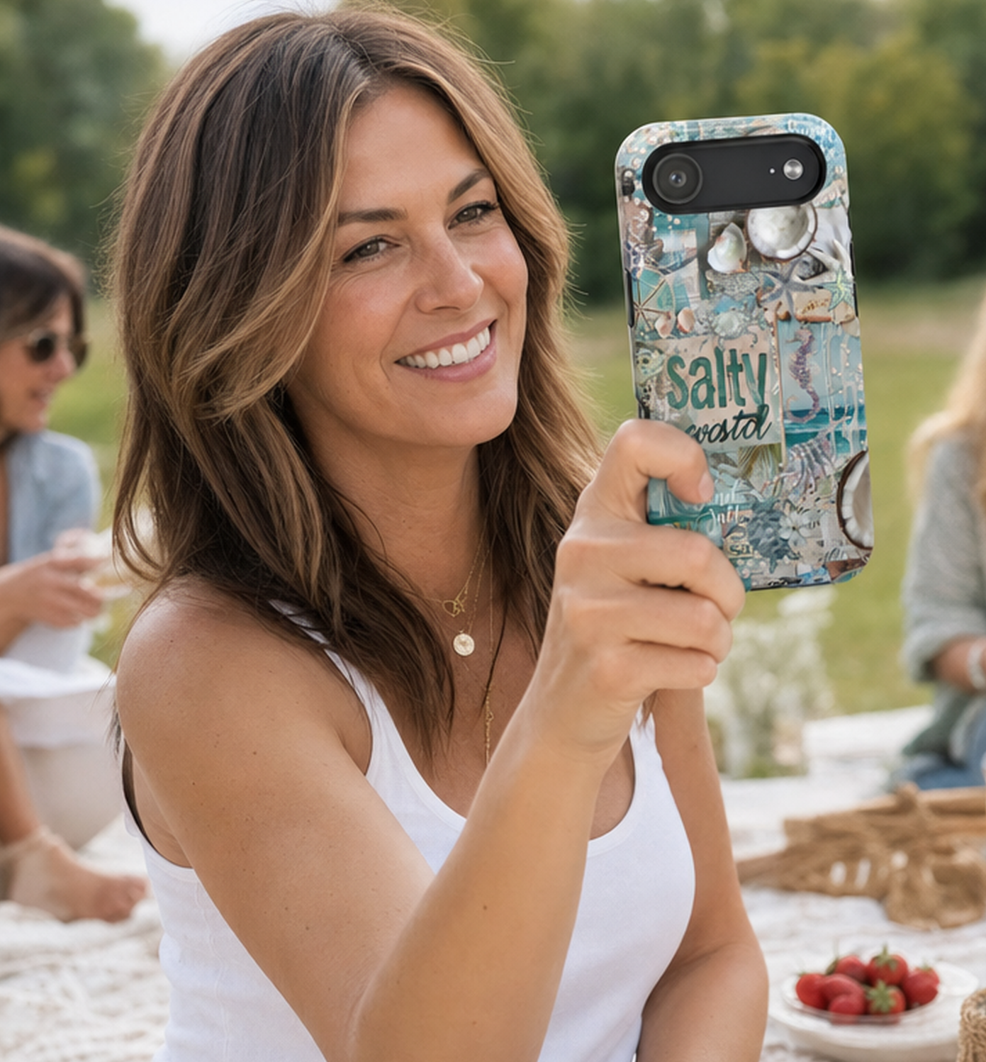 American woman in her 40s wearing a white tank top at a picnic outdoors, holding an iPhone 17 Air with a coastal themed 'salty' phone case design, by Crossroads Dandelion
