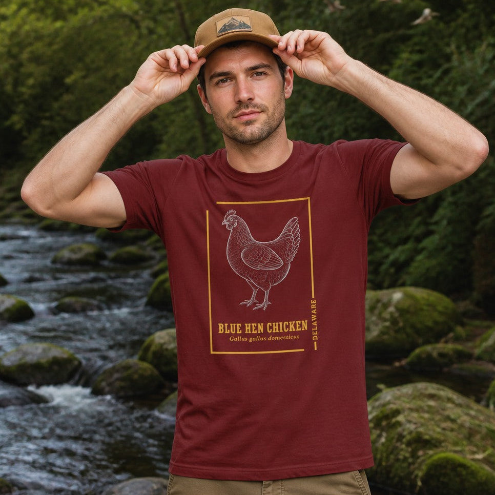 Man wearing a cardinal red Bella Canvas 3001 t-shirt with a vintage linework illustration of the Blue Hen Chicken framed in gold, cottagecore style for birdwatchers, wildlife enthusiasts, and heritage lovers, standing by a riverside in nature, by Crossroads Dandelion