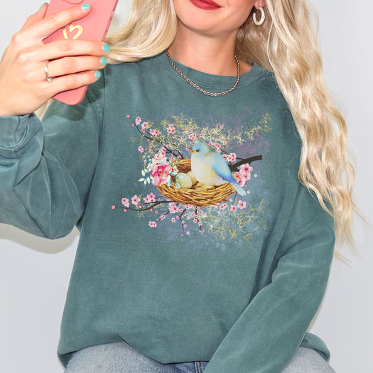 Person wearing a blue green(blue spruce color) sweatshirt with a bird and nest design, holding a phone.