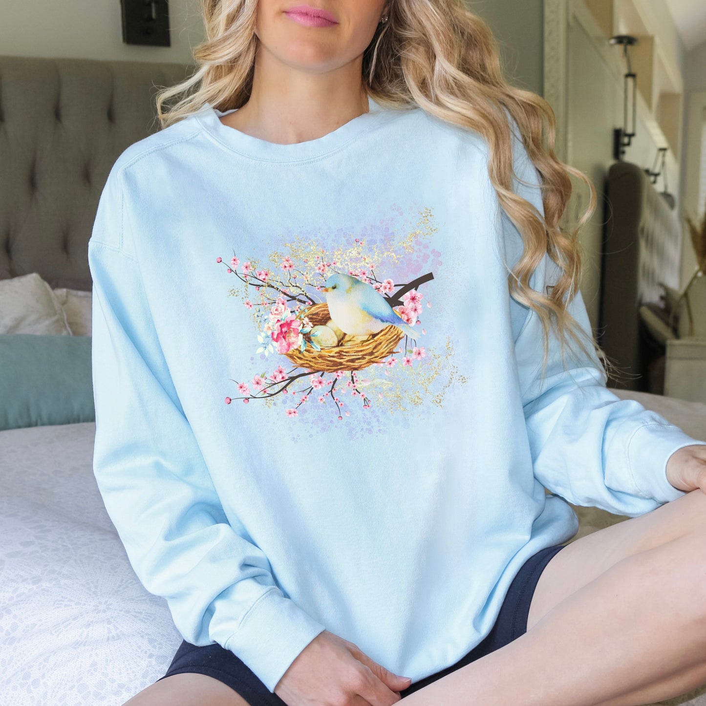 Person wearing a light blue(chambray color) sweatshirt with a bird and nest design, sitting on a bed.