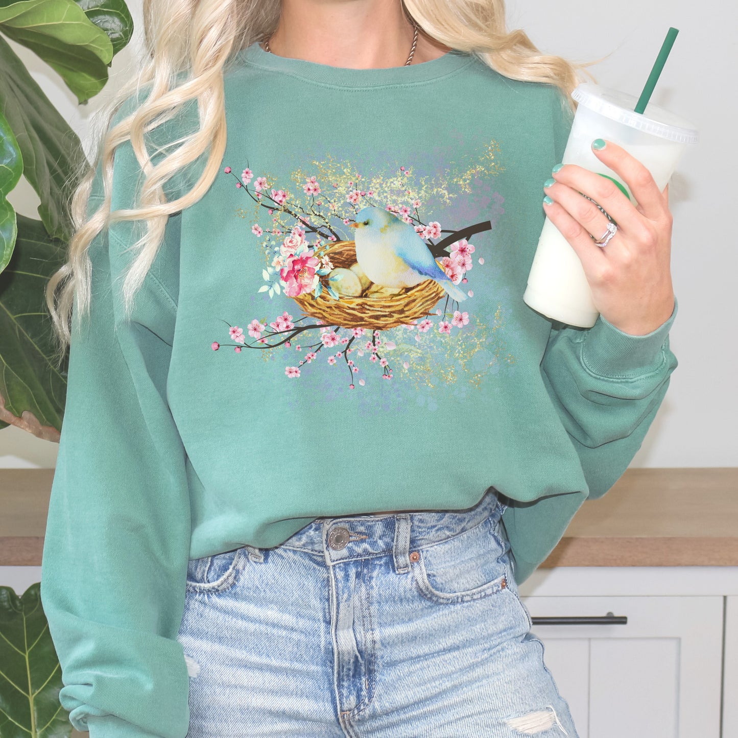Person wearing a light green sweatshirt with a bird and nest design, holding a cup.