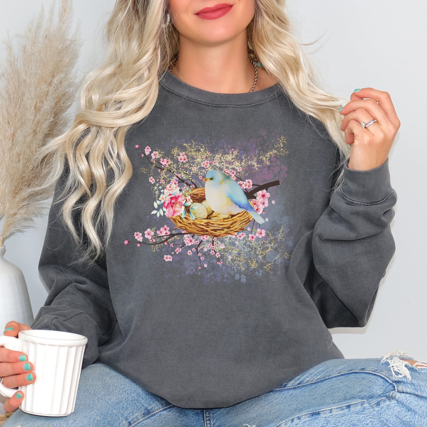 Person wearing a dark gray (pepper color) sweatshirt with a bird and nest design, holding a white mug.