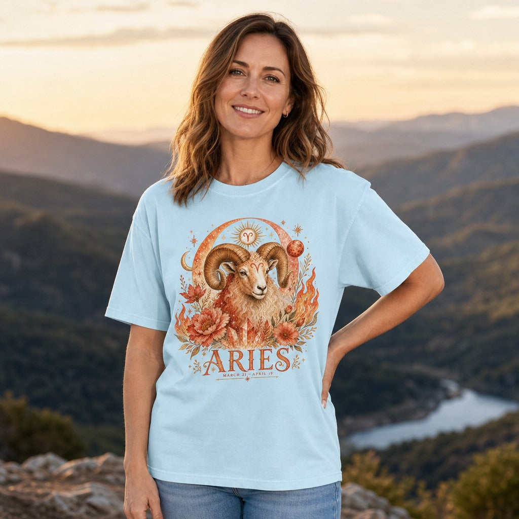 Woman in chambray blue Comfort Colors t-shirt featuring a vivid boho Aries ram surrounded by fiery florals, cosmic moons, crystals, and stars, standing in a scenic mountain setting, perfect for astrology enthusiasts, by Crossroads Dandelion