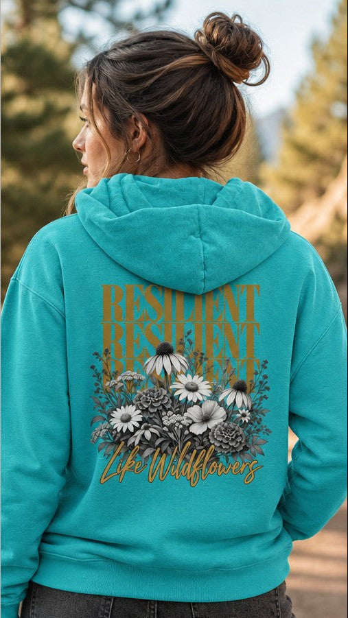 Woman wearing a seafoam hoodie with a boho wildflower and resilient life floral design, featuring daisies and foliage, outdoor setting, ideal for women who love nature-inspired apparel, by Crossroads Dandelion