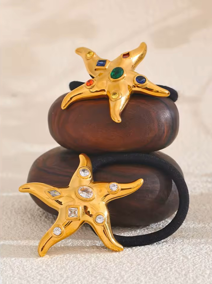 Gold starfish-shaped hair accessories with colorful and white cubic zirconia on two brown smooth rocks. 