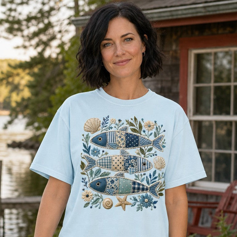 Woman in chambray Comfort Colors 1717 t-shirt featuring a cozy cottagecore coastal design with three patchwork sardine fish, embroidered shells, florals, and sea botanicals, standing outdoors by a lakeside cabin, ideal for nature-loving women, by Crossroads Dandelion