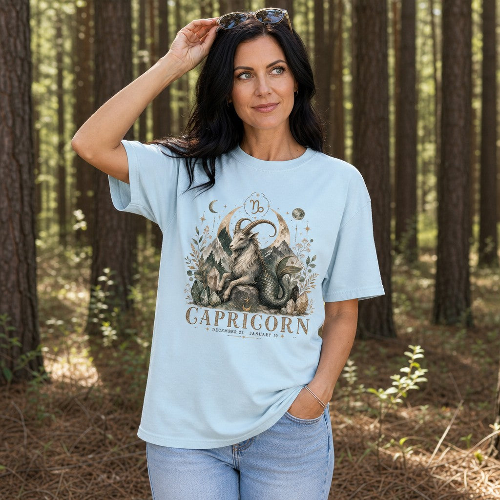 Woman wearing chambray Comfort Colors 1717 t-shirt with a vintage mystical Capricorn illustration featuring a mountain goat with mermaid tail, crescent moons, stars, mountains, botanicals, and crystals, perfect for astrology lovers, standing in a pine forest, women's zodiac tee, by Crossroads Dandelion