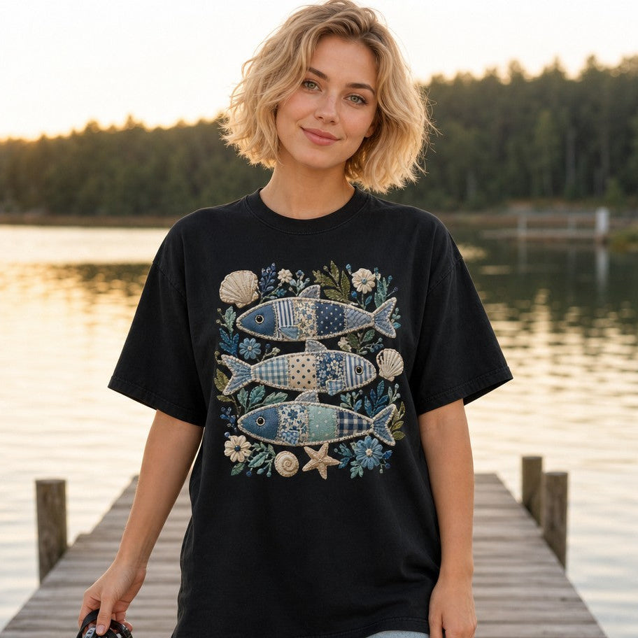 Woman wearing black Comfort Colors 1717 t-shirt featuring a cozy cottagecore and coastal patchwork sardine fish design with embroidered shells, flowers, and sea botanicals, vintage folk art style, standing on a lakeside dock, ideal for artful nature lovers and creative women, by Crossroads Dandelion