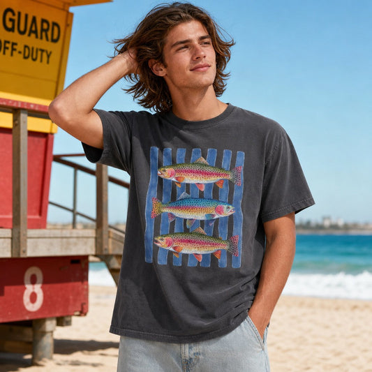 Man wearing Comfort Colors 1717 t-shirt in pepper color with hand-painted watercolor trio of rainbow trout on denim blue stripes, coastal beach setting, perfect for creekside and fishing enthusiasts, nature lovers, by Crossroads Dandelion