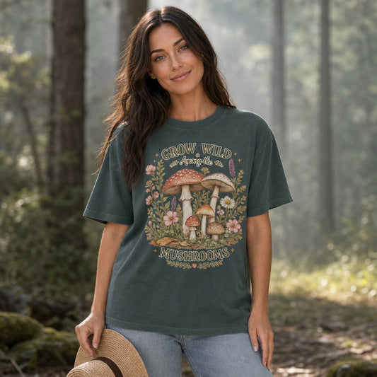 Woman in a blue spruce Comfort Colors 1717 t-shirt featuring a whimsical cottagecore design with watercolor mushrooms, wildflowers, and vintage-inspired floral details, standing in a sunlit forest, perfect for nature-loving women and mushroom enthusiasts, by Crossroads Dandelion