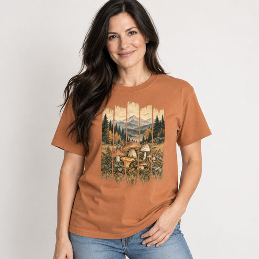 Woman wearing a yam Comfort Colors® 1717 t-shirt with a vintage folk art watercolor design of Amanita, chanterelle, and bolete mushrooms among ferns and grasses, framed by autumn trees, blue-gray mountains, and birds in a rustic barn-wood frame; perfect for foragers, gardeners, and cottagecore nature lovers, by Crossroads Dandelion