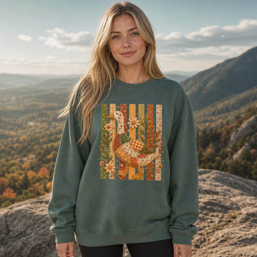 Woman in blue spruce Comfort Colors sweatshirt featuring a whimsical cottagecore patchwork goose design with floral, gingham, and polka-dot fabrics, embroidered wildflowers and leaves on a vintage folk art fence, autumn garden style, mountain overlook, perfect for nature lovers, by Crossroads Dandelion