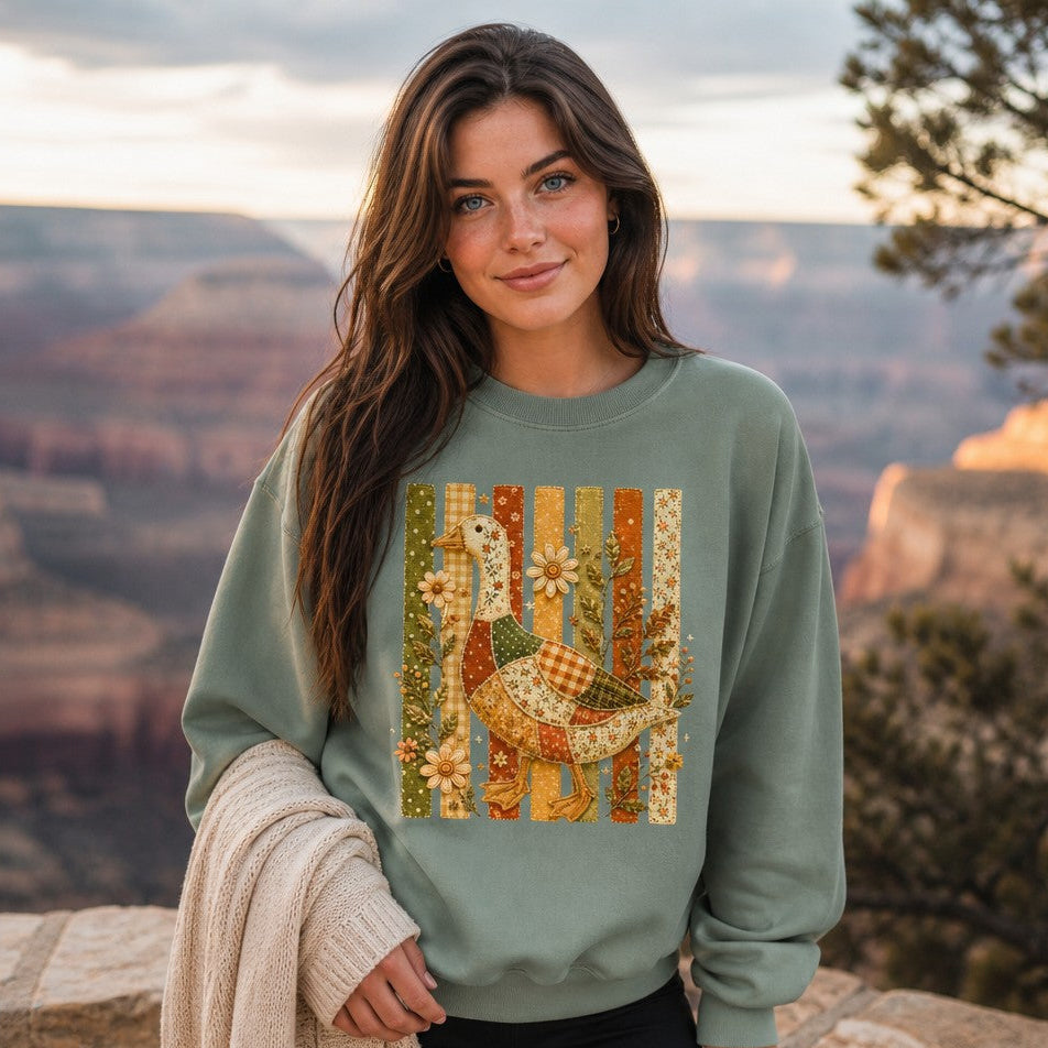 Woman wearing a light green Comfort Colors sweatshirt featuring a whimsical patchwork goose with floral, gingham, and polka-dot fabrics, vintage folk art embroidery, and stitched fence slats, photographed at the Grand Canyon, cozy autumn cottagecore nature design for women, by Crossroads Dandelion