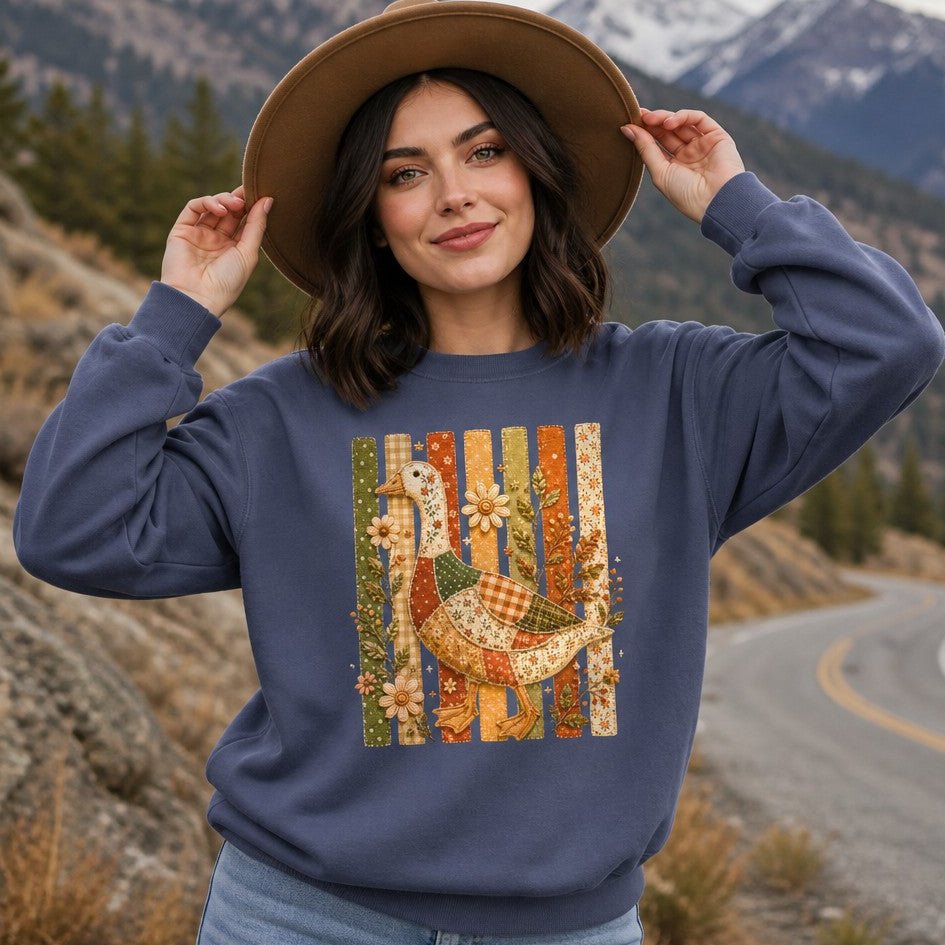 Woman wearing denim Comfort Colors 1566 sweatshirt featuring a whimsical cottagecore patchwork goose with floral, gingham, and polka-dot fabrics, embroidered wildflowers, and vintage folk art design, standing on a winding mountain road, cozy autumn aesthetic for nature and cottagecore lovers, by Crossroads Dandelion