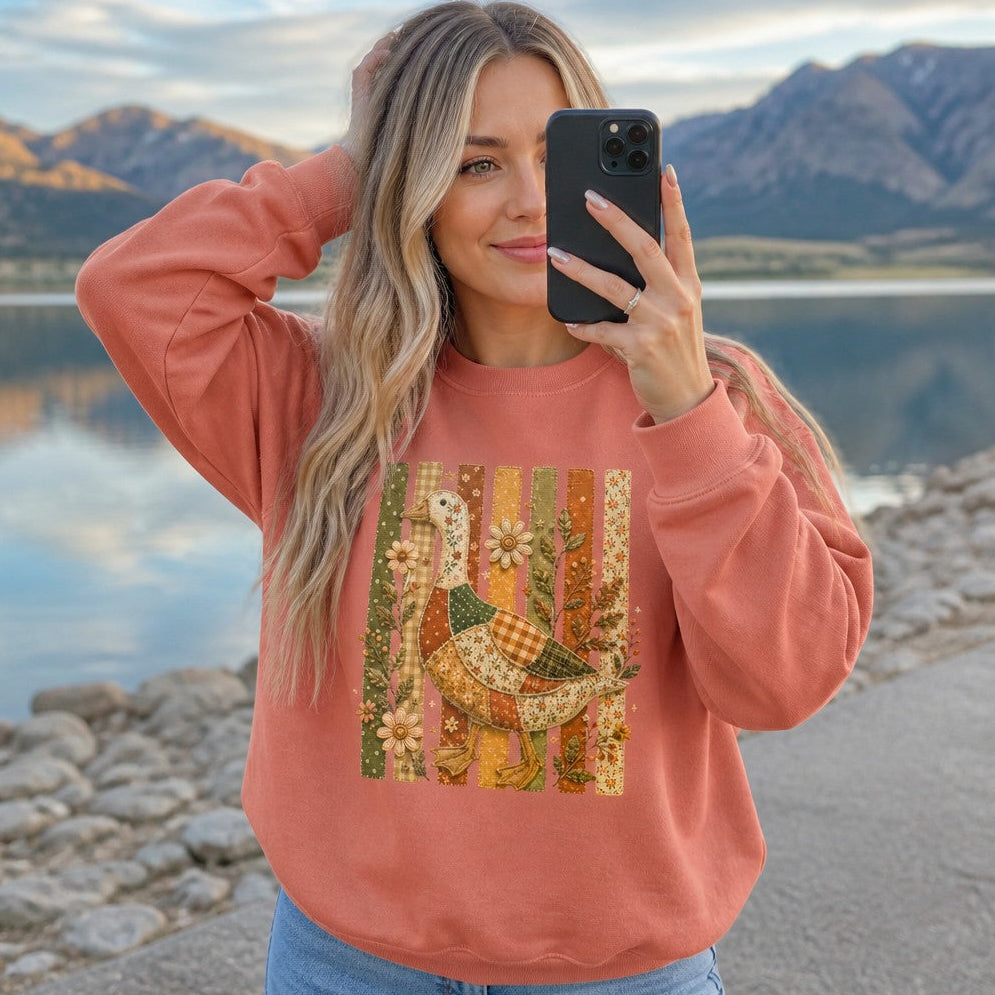 Woman wearing a terracotta Comfort Colors sweatshirt with a vintage cottagecore design featuring a whimsical patchwork goose, floral folk art, embroidered wildflowers, and autumn nature elements, standing by a scenic lakeside, cozy apparel for nature and garden lovers, by Crossroads Dandelion
