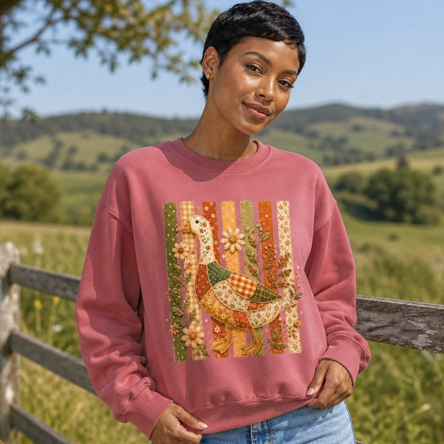 Woman wearing a crimson Comfort Colors sweatshirt featuring a whimsical patchwork goose with floral, gingham, and polka-dot fabric accents, vintage folk art and cottagecore style, embroidered wildflowers, standing by a countryside fence, perfect for nature and garden lovers, by Crossroads Dandelion