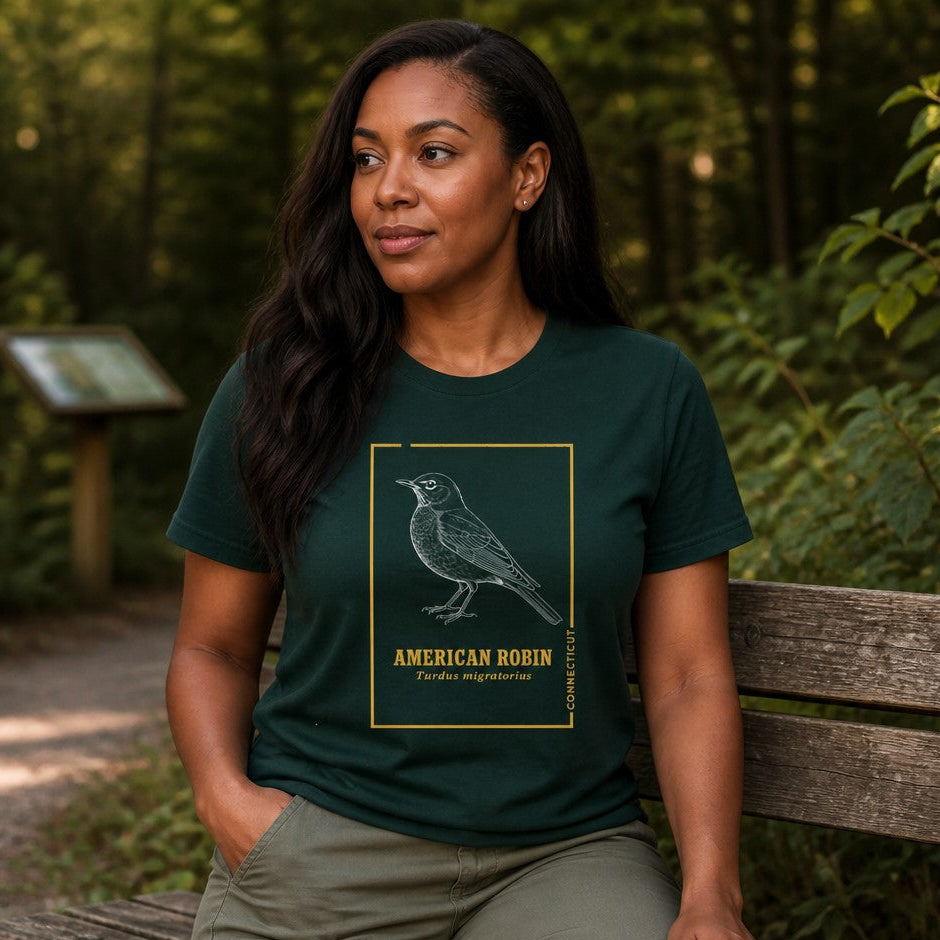 Woman wearing a forest green Bella Canvas 3001 t-shirt with a vintage cottagecore line art illustration of an American Robin, golden botanist trim, sitting on a wooded trail bench, ideal for birdwatchers and nature lovers, by Crossroads Dandelion