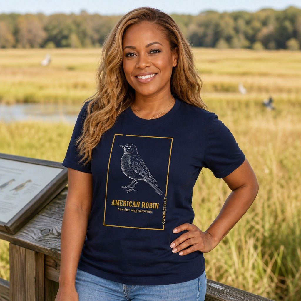 Woman wearing a navy Bella Canvas 3001 t-shirt featuring a vintage naturalist line art of an American Robin in golden botanist-inspired trim, cottagecore style, photographed on a scenic outdoor boardwalk surrounded by nature, perfect for birdwatchers and wildlife lovers, by Crossroads Dandelion