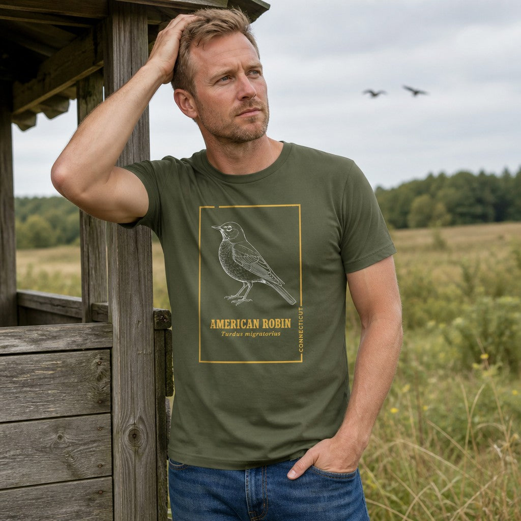 Man in military green Bella Canvas 3001 t-shirt showcasing a vintage line art American Robin design with golden botanist-inspired trim, posed by a rustic wooden shelter in a meadow, perfect for cottagecore birdwatchers and nature lovers, by Crossroads Dandelion