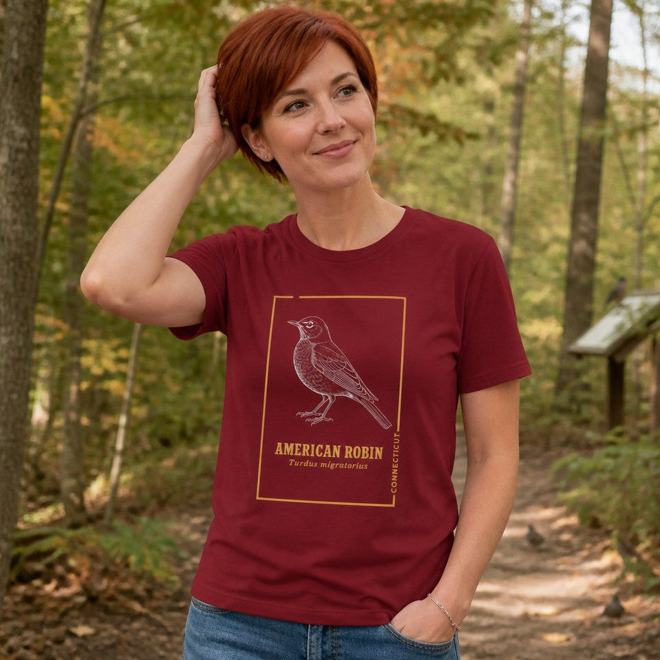 Woman wearing a cardinal red Bella Canvas 3001 t-shirt featuring a vintage naturalist line art illustration of an American Robin framed in golden botanist-inspired trim, standing on a wooded forest trail, perfect for cottagecore birdwatchers and nature lovers, by Crossroads Dandelion