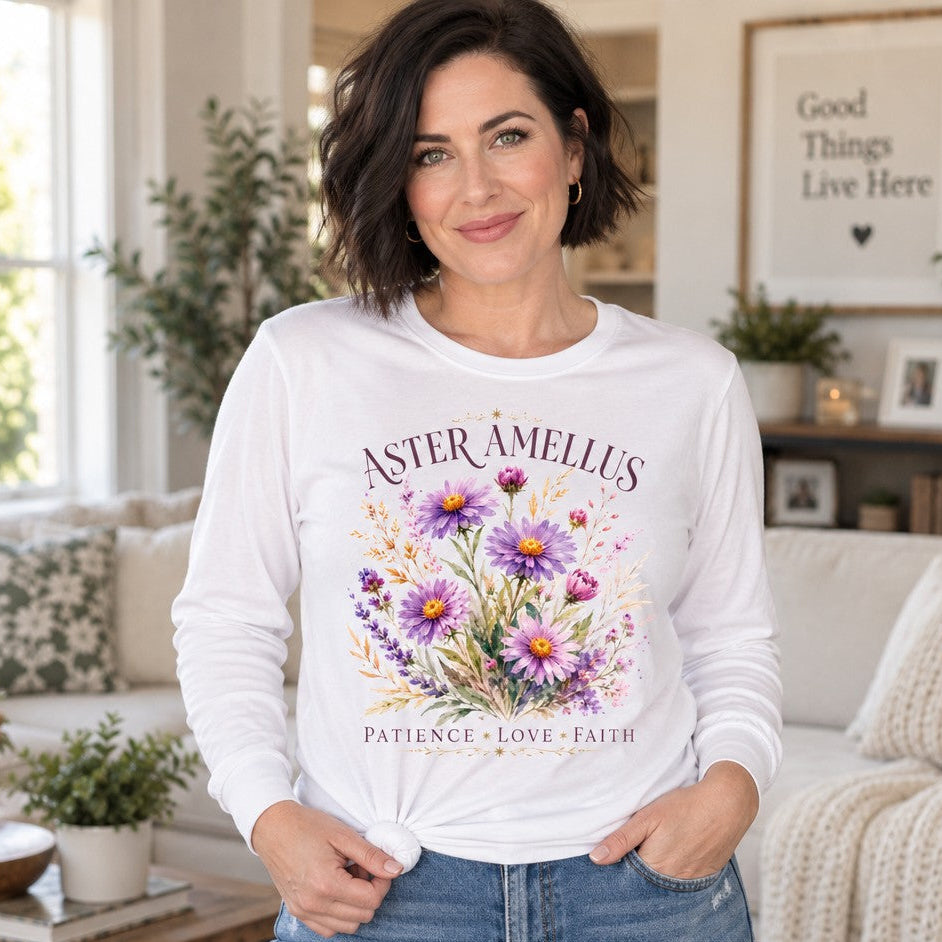Woman in a white Bella Canvas® 3501 long sleeve shirt featuring a cottagecore watercolor bouquet of purple Aster amellus with yellow centers, delicate wildflowers, and elegant serif lettering reading 'Aster Amellus Patience Love Faith', cozy living room, for September birth flower and wildflower lovers, by Crossroads Dandelion
