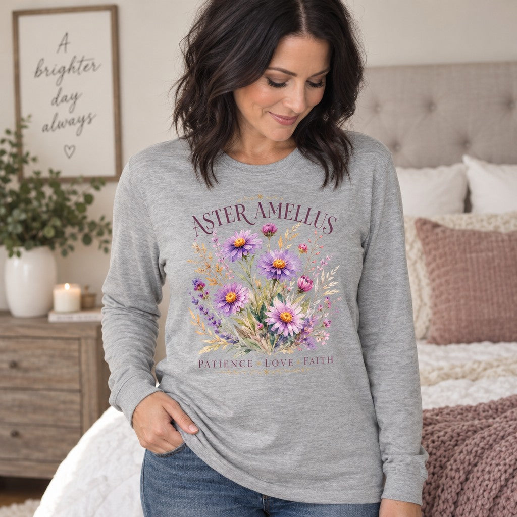 Woman in athletic heather Bella Canvas® 3501 long sleeve shirt featuring a cottagecore watercolor Aster amellus bouquet with purple, pink, and yellow blooms, elegant serif lettering, and ornamental accents, styled in a cozy bedroom setting, ideal for women and wildflower lovers, by Crossroads Dandelion