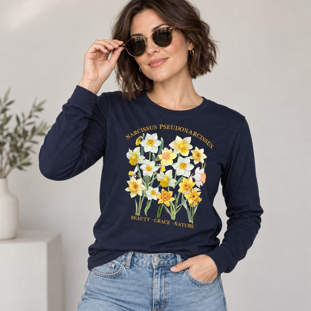 Woman wearing a navy Bella Canvas 3501 long sleeve shirt featuring a watercolor-style vintage botanical daffodil design with Latin script, perfect for creative souls and plant lovers, cottagecore aesthetic, indoor setting, by Crossroads Dandelion