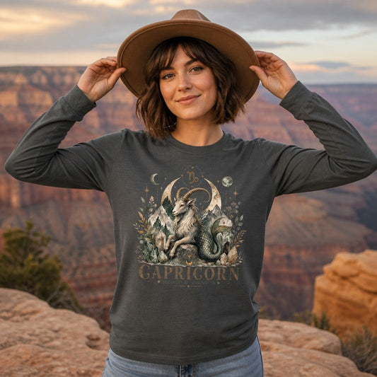Woman wearing an asphalt Bella Canvas 3501 long sleeve shirt featuring a cottagecore watercolor Capricorn sea-goat design with crystalline mountains, moon phases, and botanicals, standing at the Grand Canyon, astrology and zodiac lovers apparel, by Crossroads Dandelion