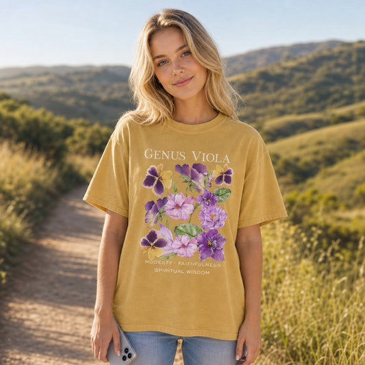 Woman wearing mustard Comfort Colors 1717 t-shirt featuring a vintage cottagecore watercolor illustration of violet and pansy flowers from the genus Viola, botanical floral design, casual outdoor nature walk setting, for women and floral nature lovers, by Crossroads Dandelion