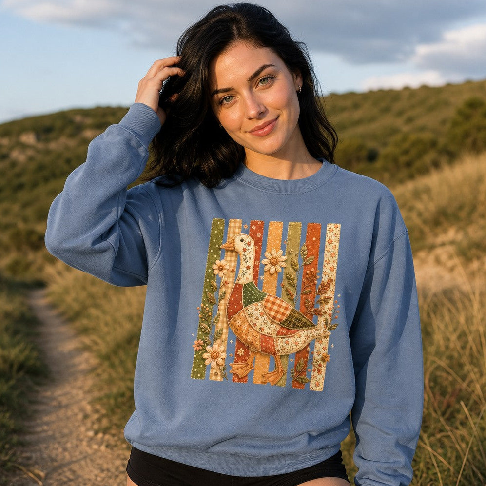 Woman wearing a blue jean Comfort Colors sweatshirt featuring a whimsical patchwork goose with floral, gingham, and polka-dot fabrics, set against pastel stitched slats and wildflowers in a vintage folk art style, outdoors in a grassy hillside setting, cottagecore and nature lovers, by Crossroads Dandelion
