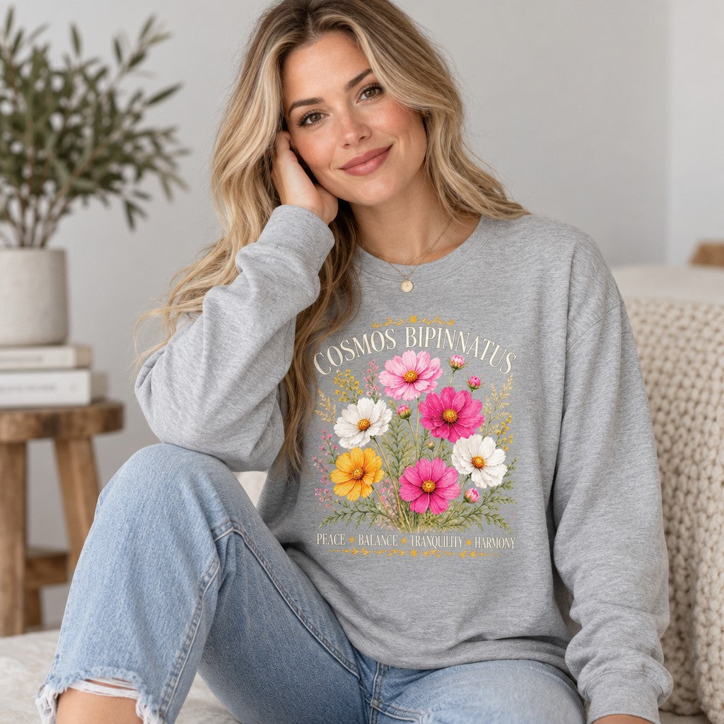 Woman in athletic heather Bella Canvas 3501 long sleeve featuring a vintage watercolor botanical illustration of cosmos bipinnatus flowers in soft pink, white, and yellow with golden celestial accents, cozy indoor setting, ideal for cottagecore fans and October birthdays, by Crossroads Dandelion