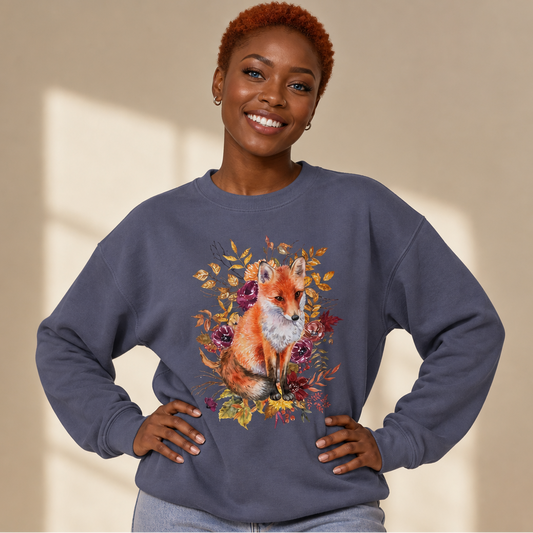 A smiling woman with short red hair wears a denim blue color crewneck sweatshirt featuring a detailed graphic of a fox surrounded by autumn leaves. She is posing with her hands on her hips against a warm beige background with soft window light shadows.