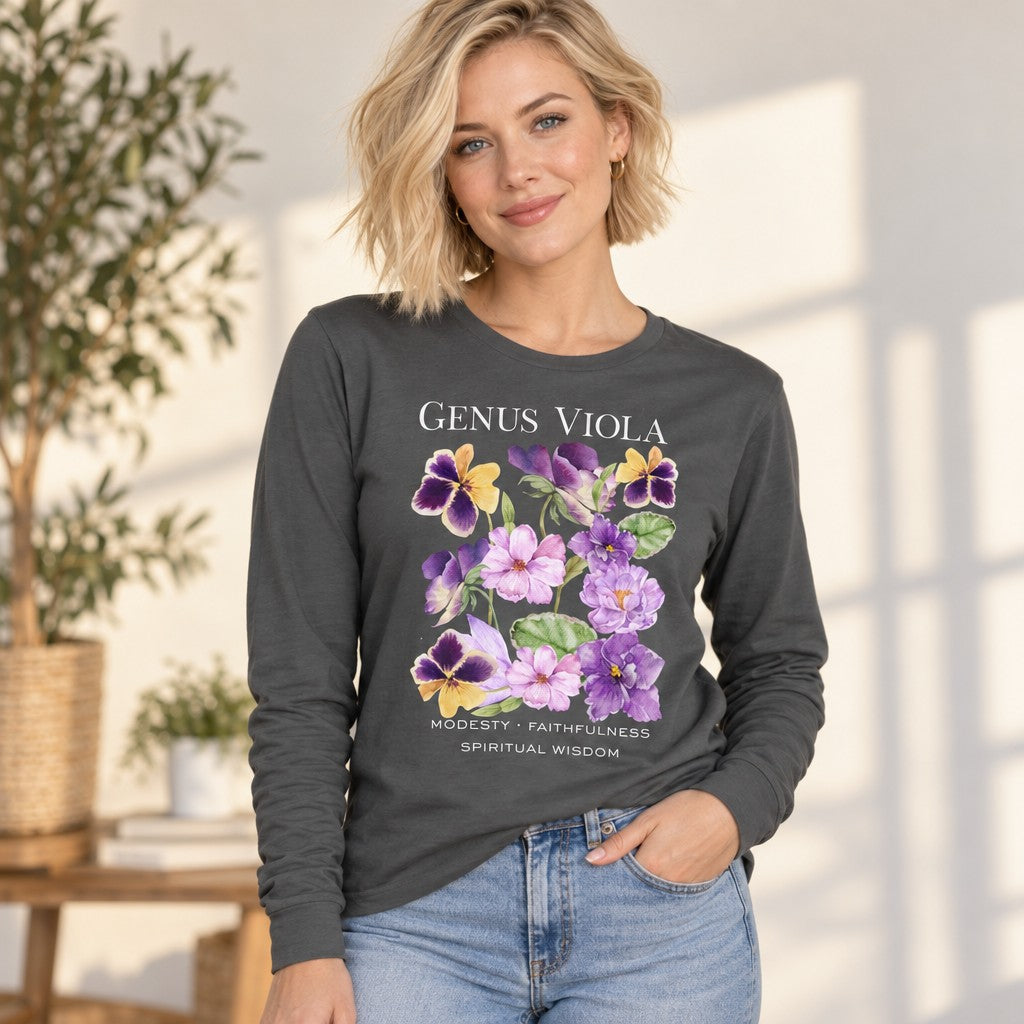 Blonde woman wearing an asphalt Bella Canvas 3501 long sleeve featuring a lush watercolor illustration of violets, pansies, and heart-shaped leaves with hand-lettered botanical meanings, cottagecore floral vintage nature journal design, relaxing in a cozy sunlit home, perfect for botanical art lovers, by Crossroads Dandelion