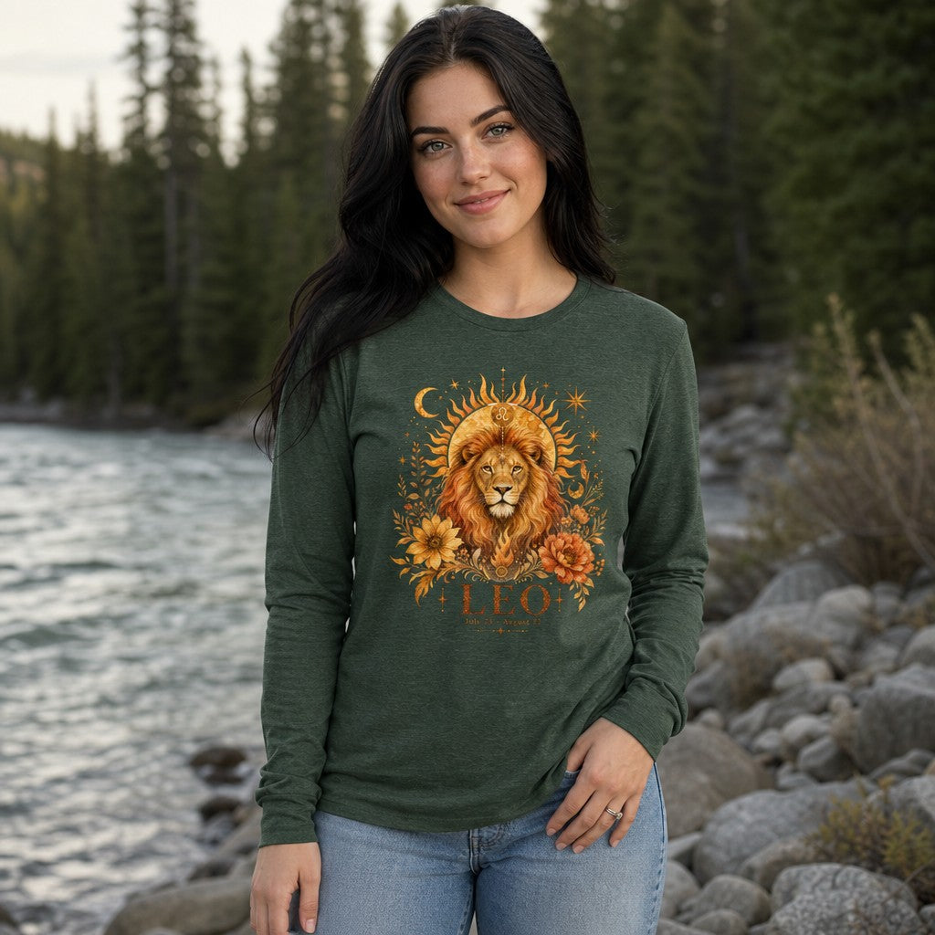 Woman wearing a heather forest Bella Canvas 3501 long sleeve shirt featuring a hand-painted cottagecore Leo lion zodiac design with sunbeams, moons, stars, and lush florals, standing by a river in a pine forest, perfect for astrology lovers and botanical art fans, by Crossroads Dandelion