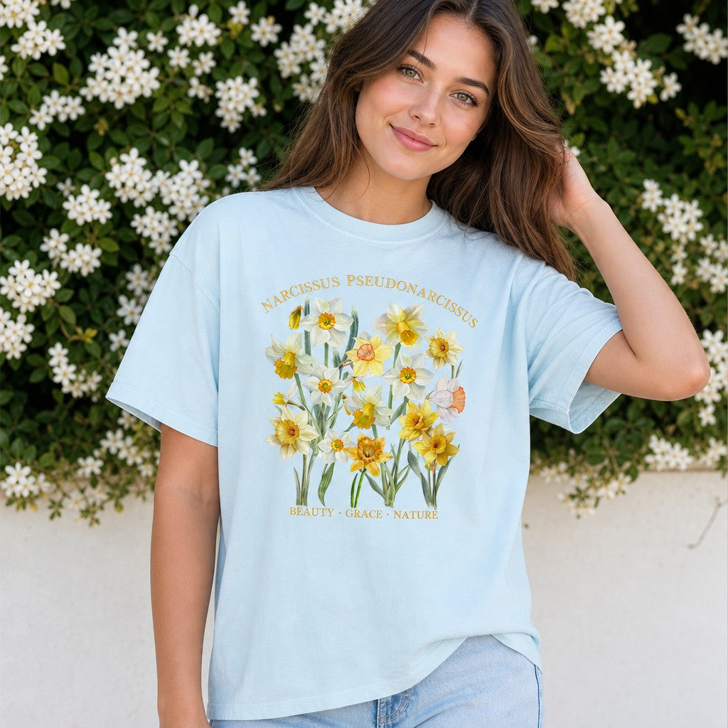 Woman in chambray Comfort Colors 1717 t-shirt featuring a vintage cottagecore watercolor illustration of Narcissus pseudonarcissus daffodils with golden botanical text, floral labels, and a serene garden background, ideal for floral enthusiasts and women, by Crossroads Dandelion
