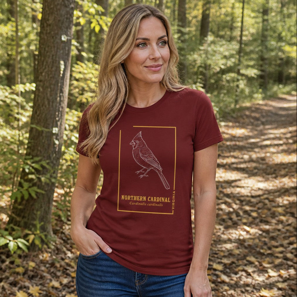 Woman wearing a cardinal red Bella Canvas 3001 t-shirt featuring a white hand-drawn northern cardinal in vintage-style gold frame and elegant serif type, cottagecore botanical line art design, walking in a sunlit forest, perfect for birdwatchers and nature lovers, by Crossroads Dandelion