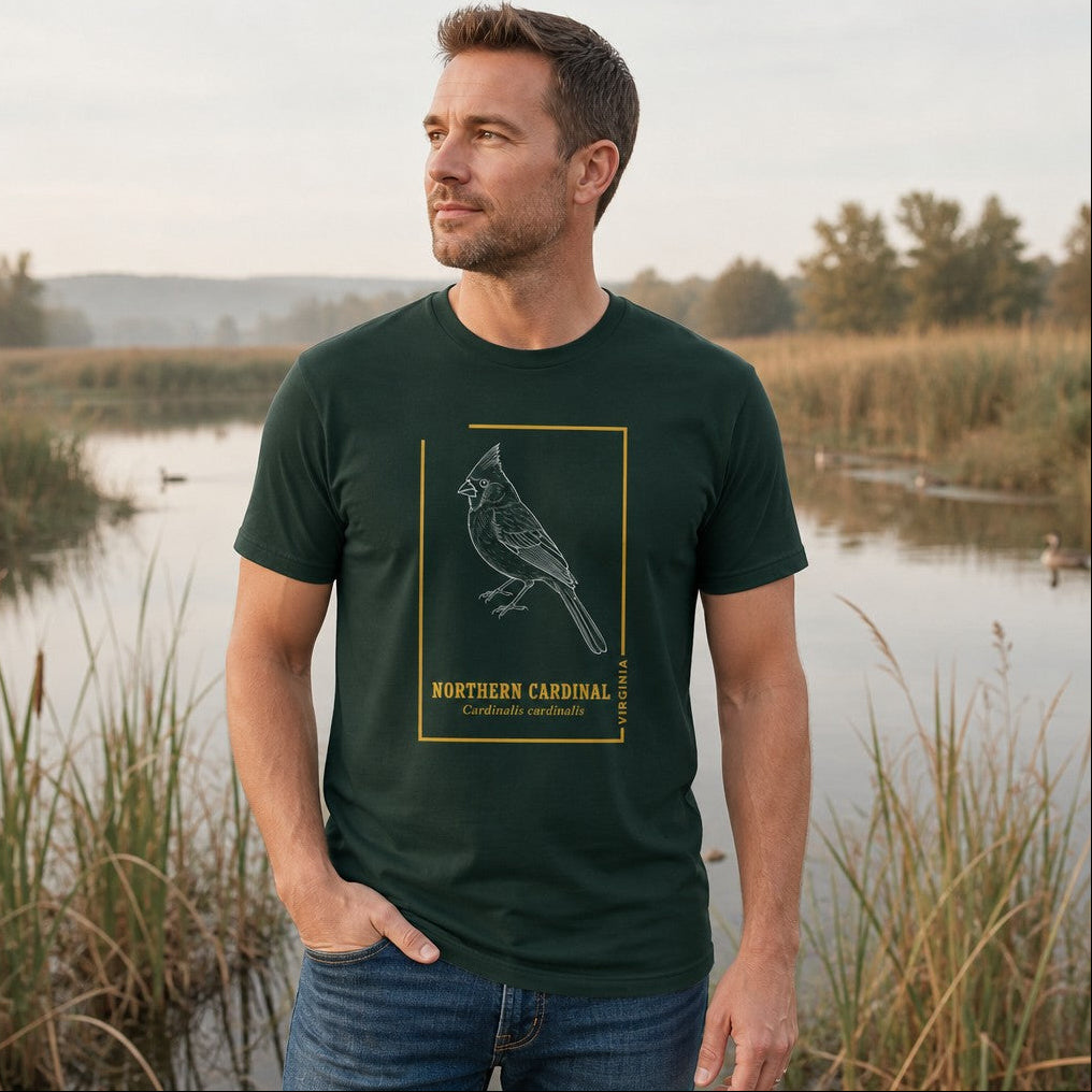 Man wearing a forest green Bella Canvas 3001 t-shirt featuring a white hand-drawn line art northern cardinal framed in gold vintage style accents with serif typography, standing outdoors by a pond, cottagecore and vintage birdwatcher gift, by Crossroads Dandelion