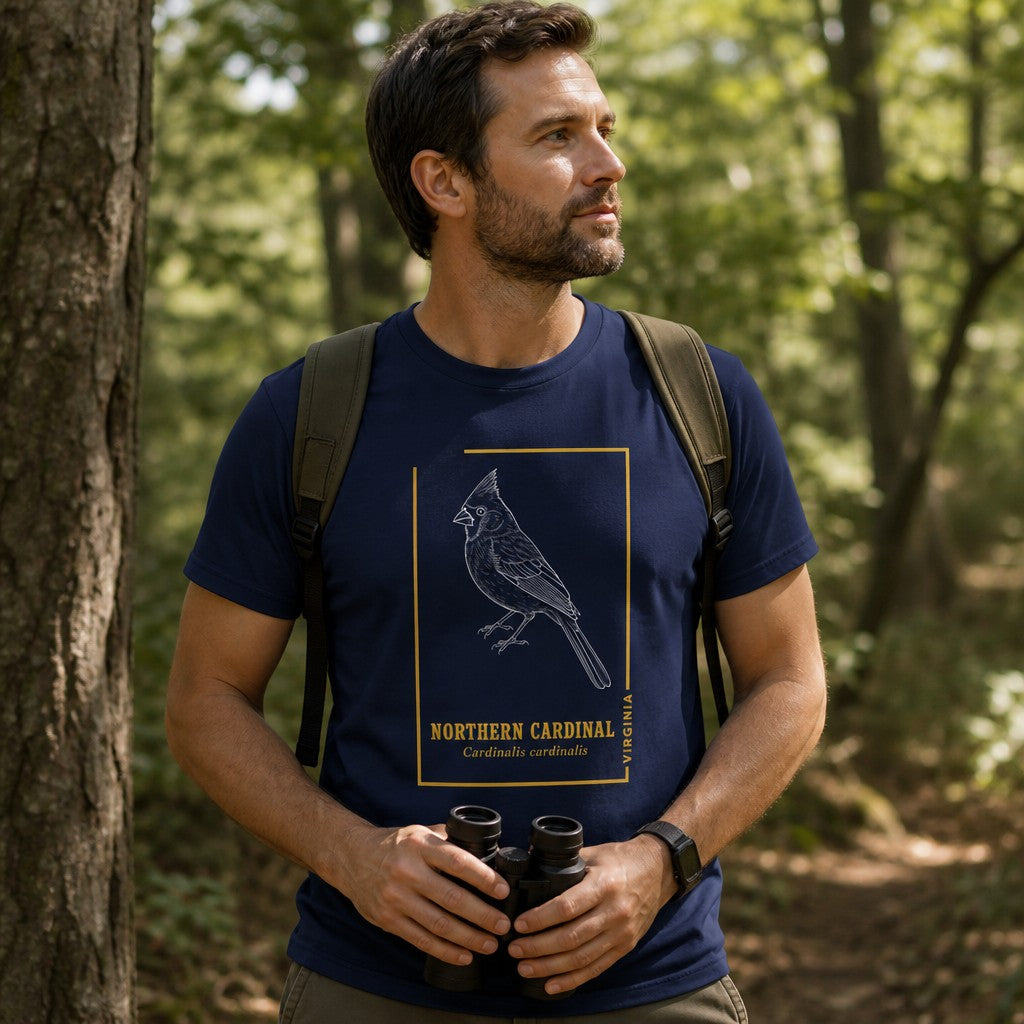 Man in navy Bella Canvas 3001 t-shirt with hand-drawn white line art illustration of a northern cardinal, vintage gold accents and serif text, standing in a sunlit forest, ideal for birdwatchers and nature lovers, by Crossroads Dandelion