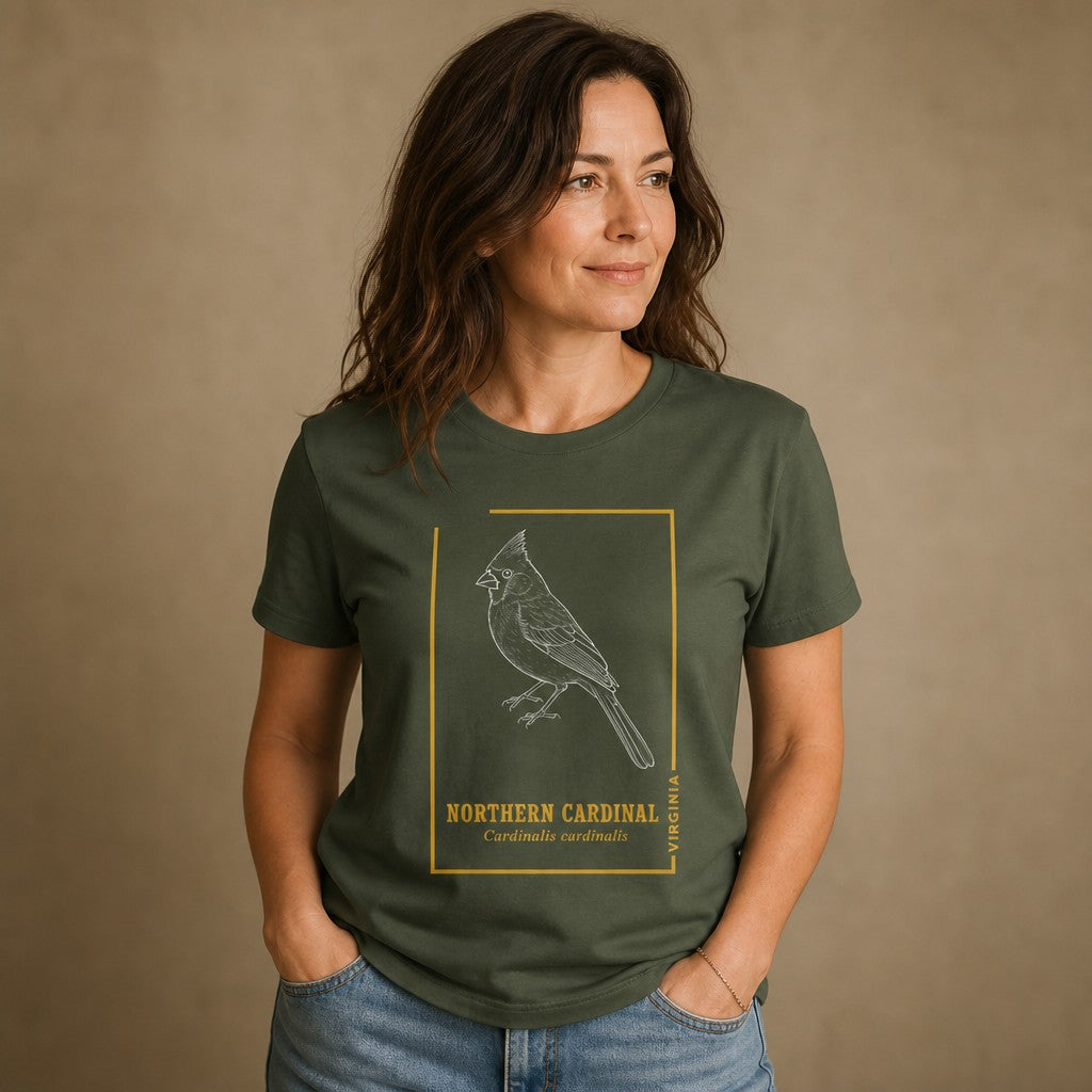 Woman in military green Bella Canvas 3001 t-shirt with a white line art illustration of a northern cardinal, framed in vintage gold accents, cottagecore style, for birdwatchers and botanical print lovers, studio background, by Crossroads Dandelion