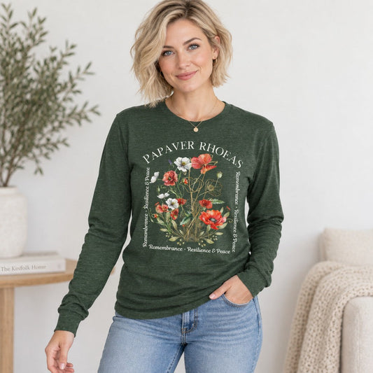 Woman in heather forest green Bella Canvas long sleeve shirt featuring a lush watercolor poppy floral design with golden botanical linework and poetic text, cozy home cottagecore scene, perfect for nature and art lovers, by Crossroads Dandelion
