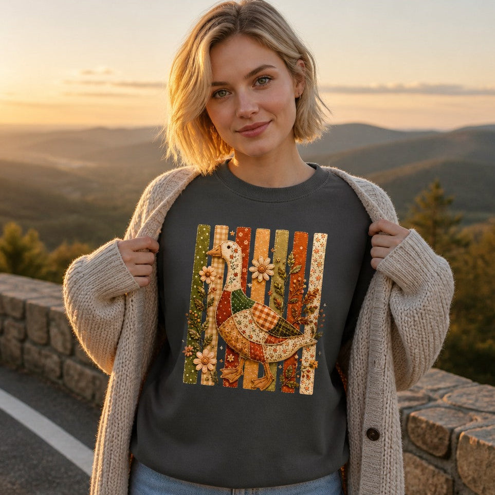 Blonde woman in a pepper Comfort Colors 1566 sweatshirt featuring a whimsical patchwork goose made from floral, gingham, and polka-dot patterns, set against vintage stitched fence slats with embroidered wildflowers; autumn mountain view, cozy cottagecore style for nature and folk art lovers, by Crossroads Dandelion
