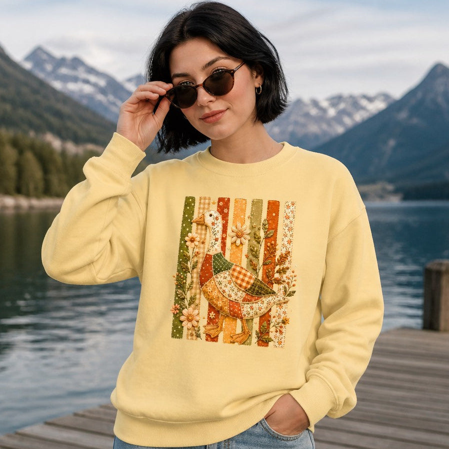 Woman wearing a butter yellow Comfort Colors 1566 sweatshirt featuring a whimsical patchwork goose with floral, gingham, and polka-dot fabrics, embroidered wildflowers, vintage folk art cottagecore style, posed by a scenic mountain lake, cozy autumn garden lovers, by Crossroads Dandelion