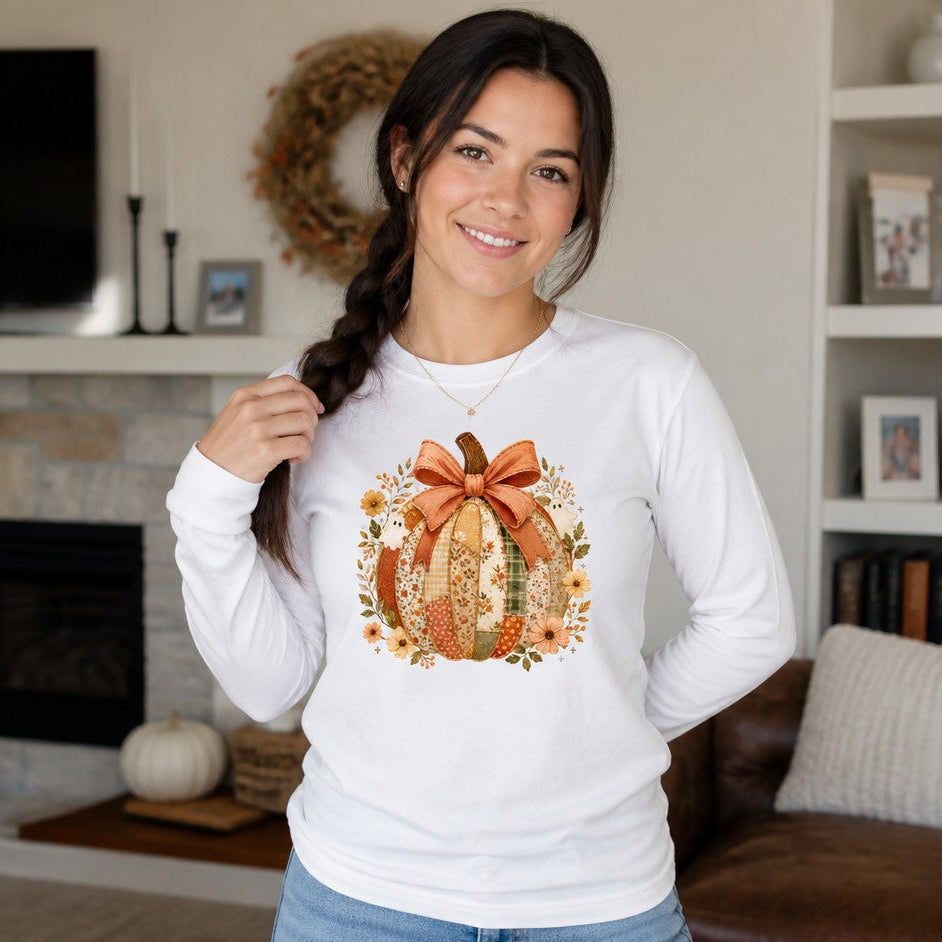 Woman wearing a white Bella Canvas 3501 long sleeve shirt with a whimsical cottagecore design featuring a patchwork pumpkin, faded coral bow, and vintage-style ghosts with sage and russet wildflowers, cozy autumn living room scene, perfect for cottagecore and fall fashion lovers, by Crossroads Dandelion